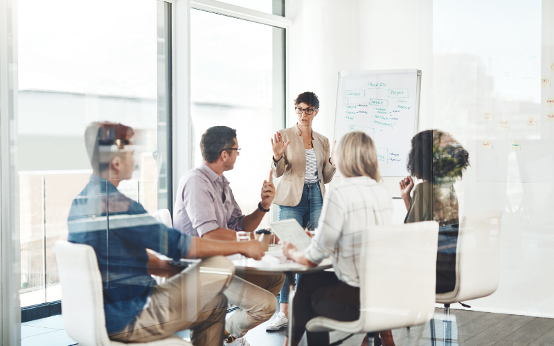 Multi-coloured graphic illustration of a group of people standing together, listening as a facilitator gives a presentation. There are posters on the wall as part of the facilitator's presentation.